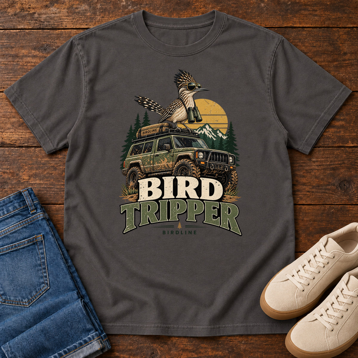 Graphite Bird Tripper T-Shirt with a crested, long-tailed bird with binoculars above an off-road vehicle and Bird Tripper lettering, front flat lay on wood.