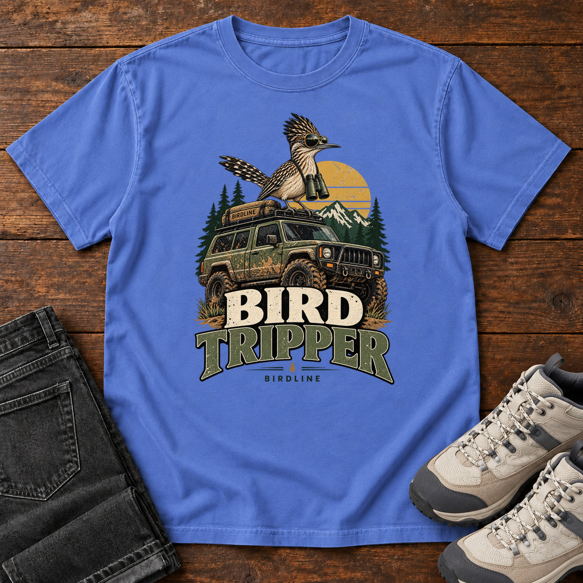 Mystic Blue Bird Tripper T-Shirt with a crested, long-tailed bird with binoculars above an off-road vehicle and Bird Tripper lettering, front flat lay on wood.