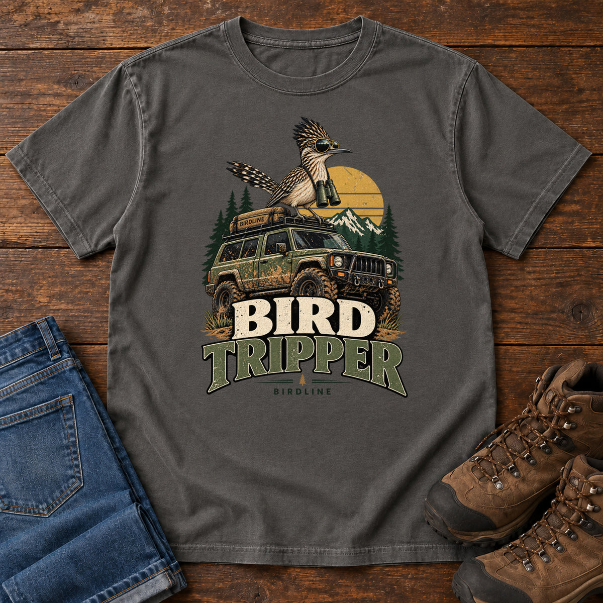 Pepper Bird Tripper T-Shirt with a crested, long-tailed bird with binoculars above an off-road vehicle and Bird Tripper lettering, front flat lay on wood.