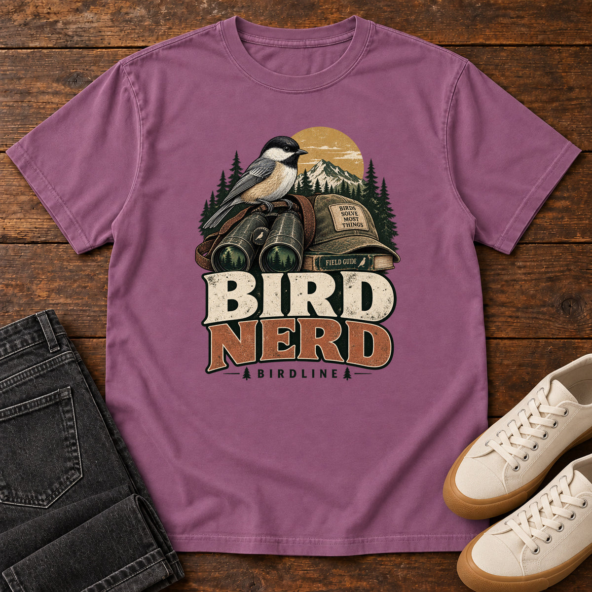 Bird Nerd Binoculars T-Shirt in Berry, front laid flat on wood, featuring a black-capped, cream-and-gray bird above binoculars and a cap, with mountains and bold lettering.