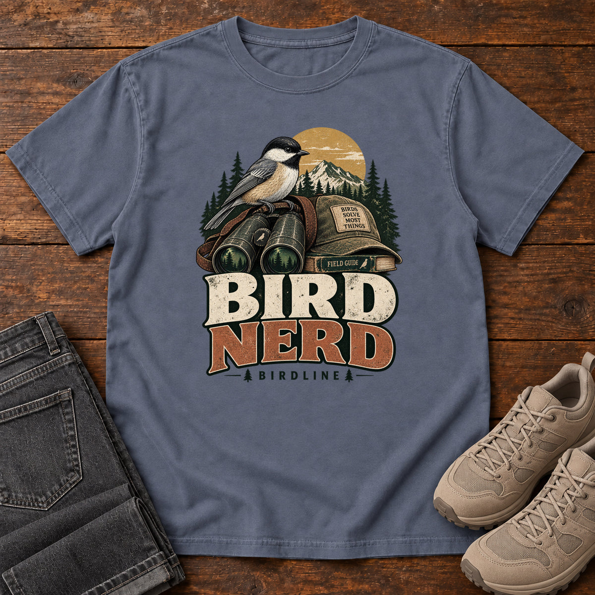 Bird Nerd Binoculars T-Shirt in Denim, front laid flat on wood, featuring a black-capped, cream-and-gray bird above binoculars and a cap, with mountains and bold lettering.