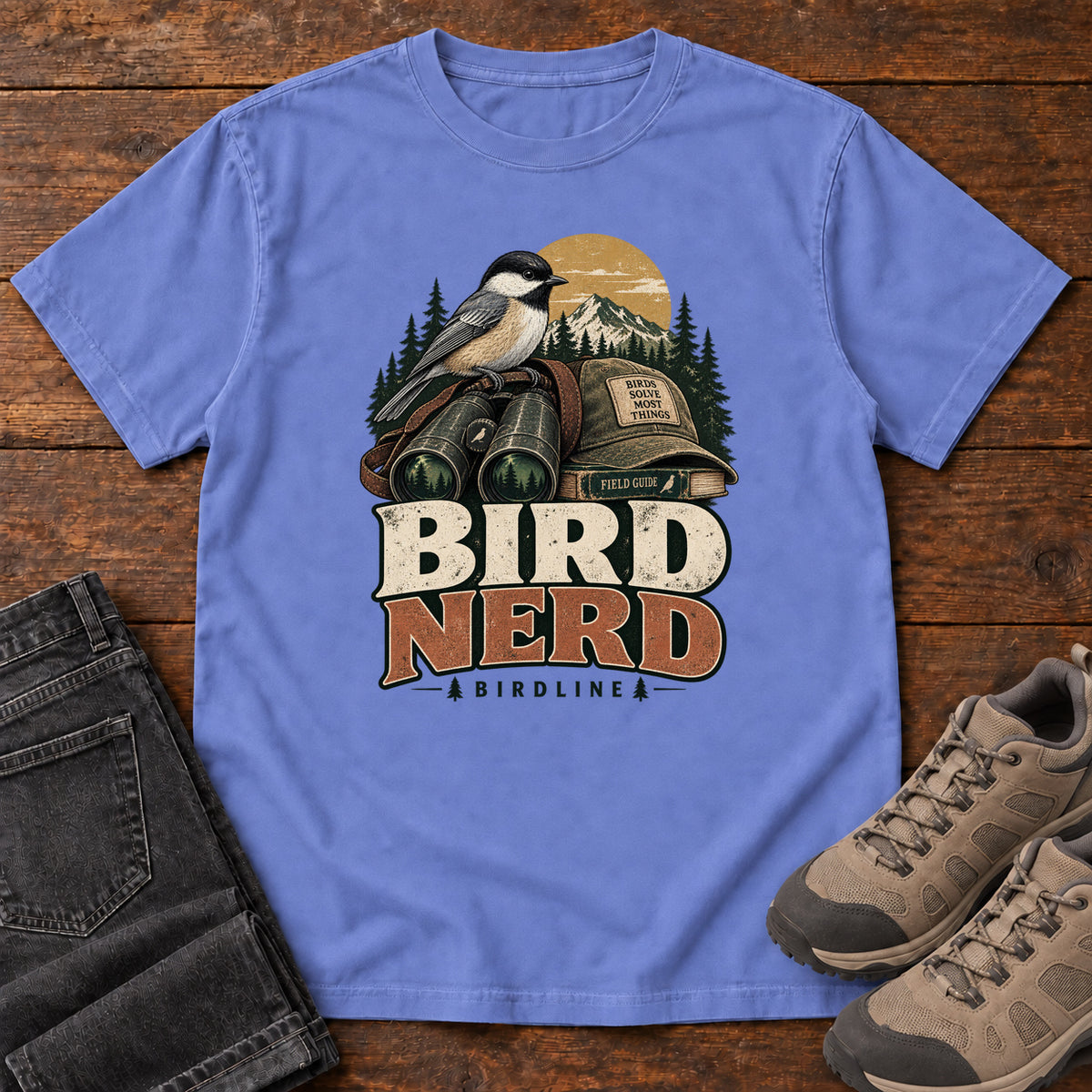 Bird Nerd Binoculars T-Shirt in Flo Blue, front laid flat on wood, featuring a black-capped, cream-and-gray bird above binoculars and a cap, with mountains and bold lettering.