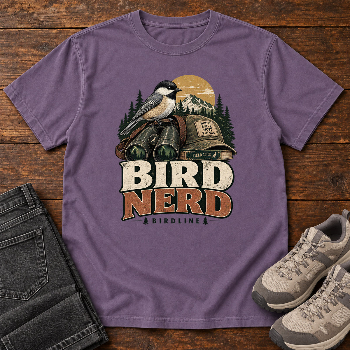 Bird Nerd Binoculars T-Shirt in Grape, front laid flat on wood, featuring a black-capped, cream-and-gray bird above binoculars and a cap, with mountains and bold lettering.