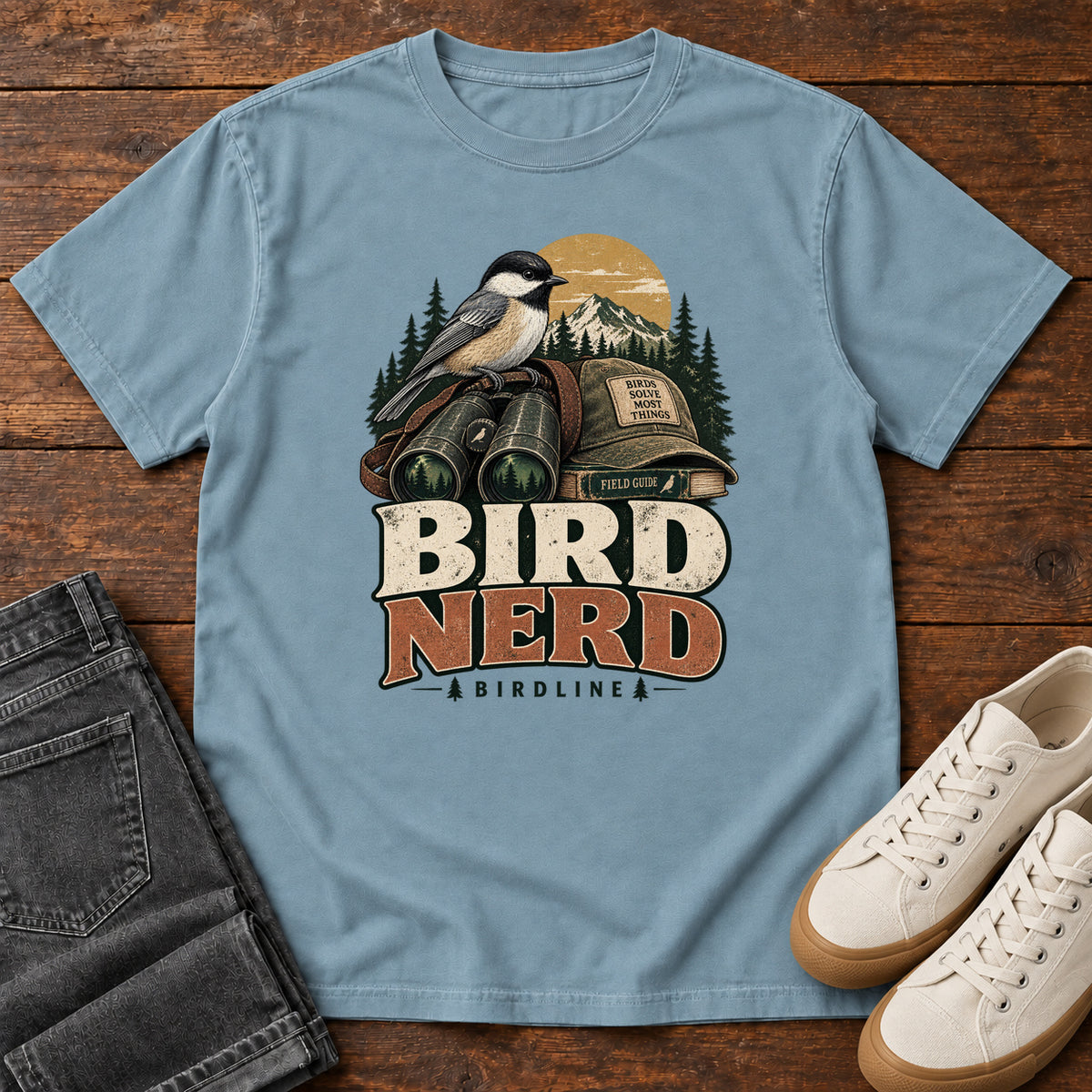 Bird Nerd Binoculars T-Shirt in Ice Blue, front laid flat on wood, featuring a black-capped, cream-and-gray bird above binoculars and a cap, with mountains and bold lettering.