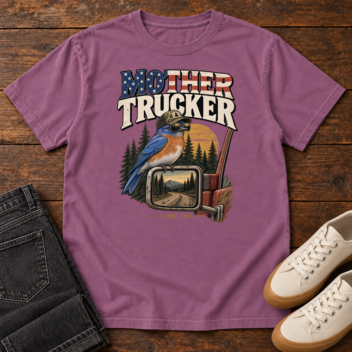 Berry Mother Trucker Side Mirror T-Shirt with a blue-and-orange bird perched on a truck mirror beneath flag-patterned lettering, front flat lay on wood.