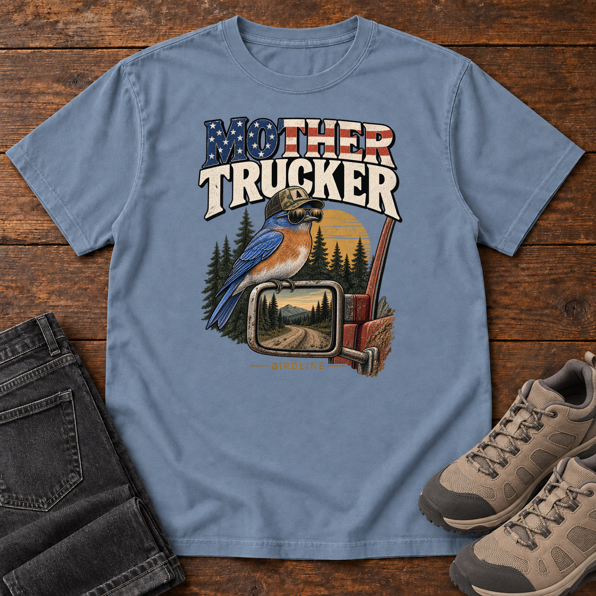 Blue Jean Mother Trucker Side Mirror T-Shirt with a blue-and-orange bird perched on a truck mirror beneath flag-patterned lettering, front flat lay on wood.