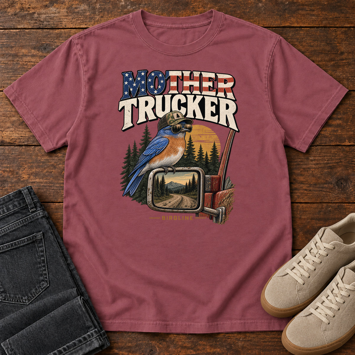 Brick Mother Trucker Side Mirror T-Shirt with a blue-and-orange bird perched on a truck mirror beneath flag-patterned lettering, front flat lay on wood.