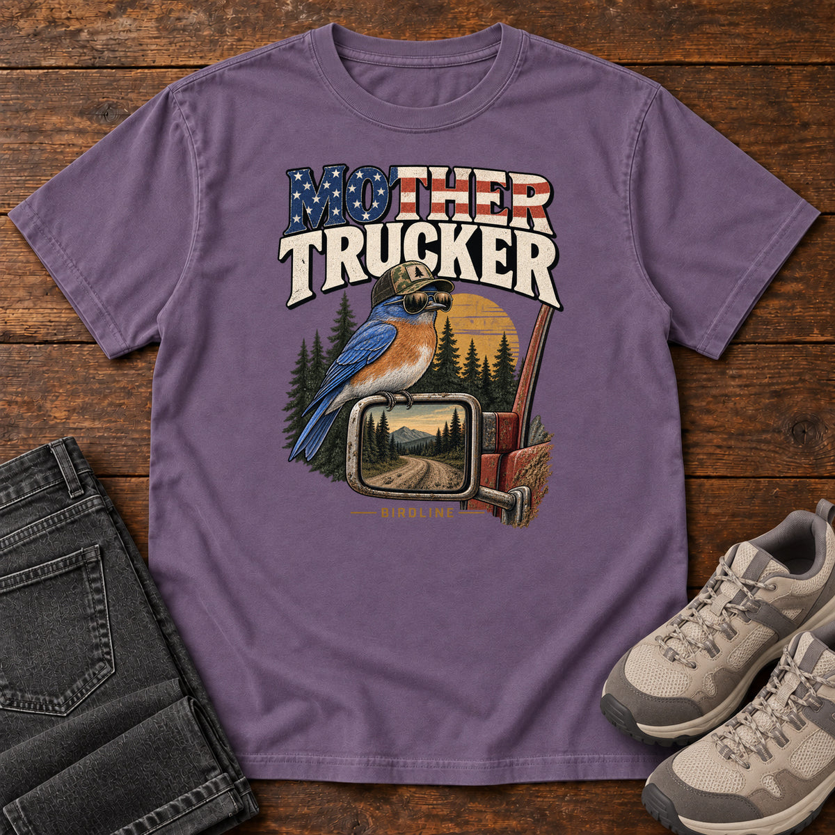 Grape Mother Trucker Side Mirror T-Shirt with a blue-and-orange bird perched on a truck mirror beneath flag-patterned lettering, front flat lay on wood.