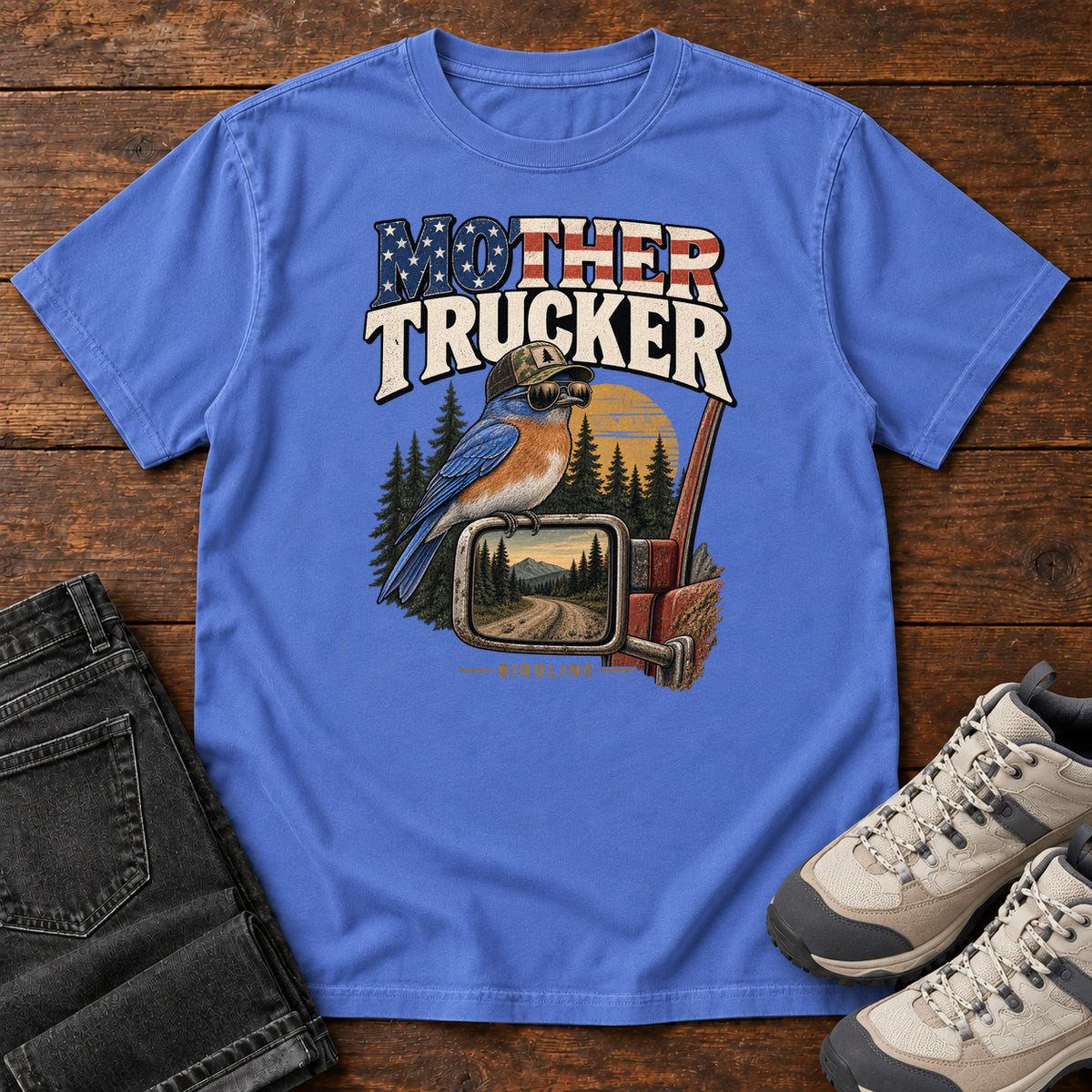 Mystic Blue Mother Trucker Side Mirror T-Shirt with a blue-and-orange bird perched on a truck mirror beneath flag-patterned lettering, front flat lay on wood.