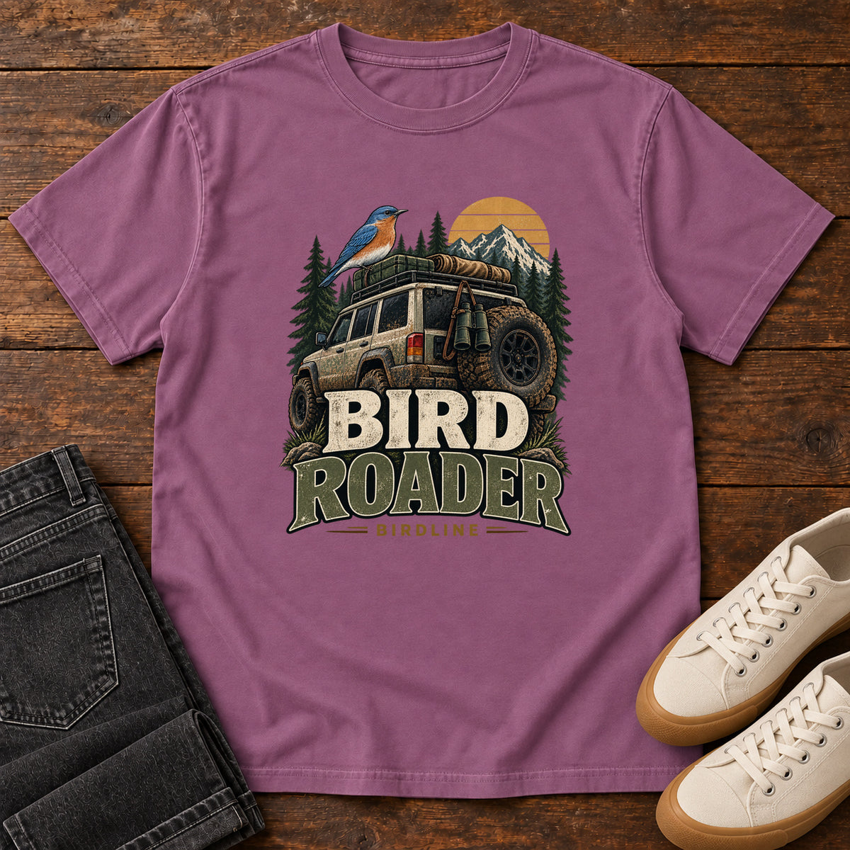Berry Bird Roader Tailgate T-Shirt with a bird on an off-road vehicle beneath mountains and a golden sun; front flat lay on wood.