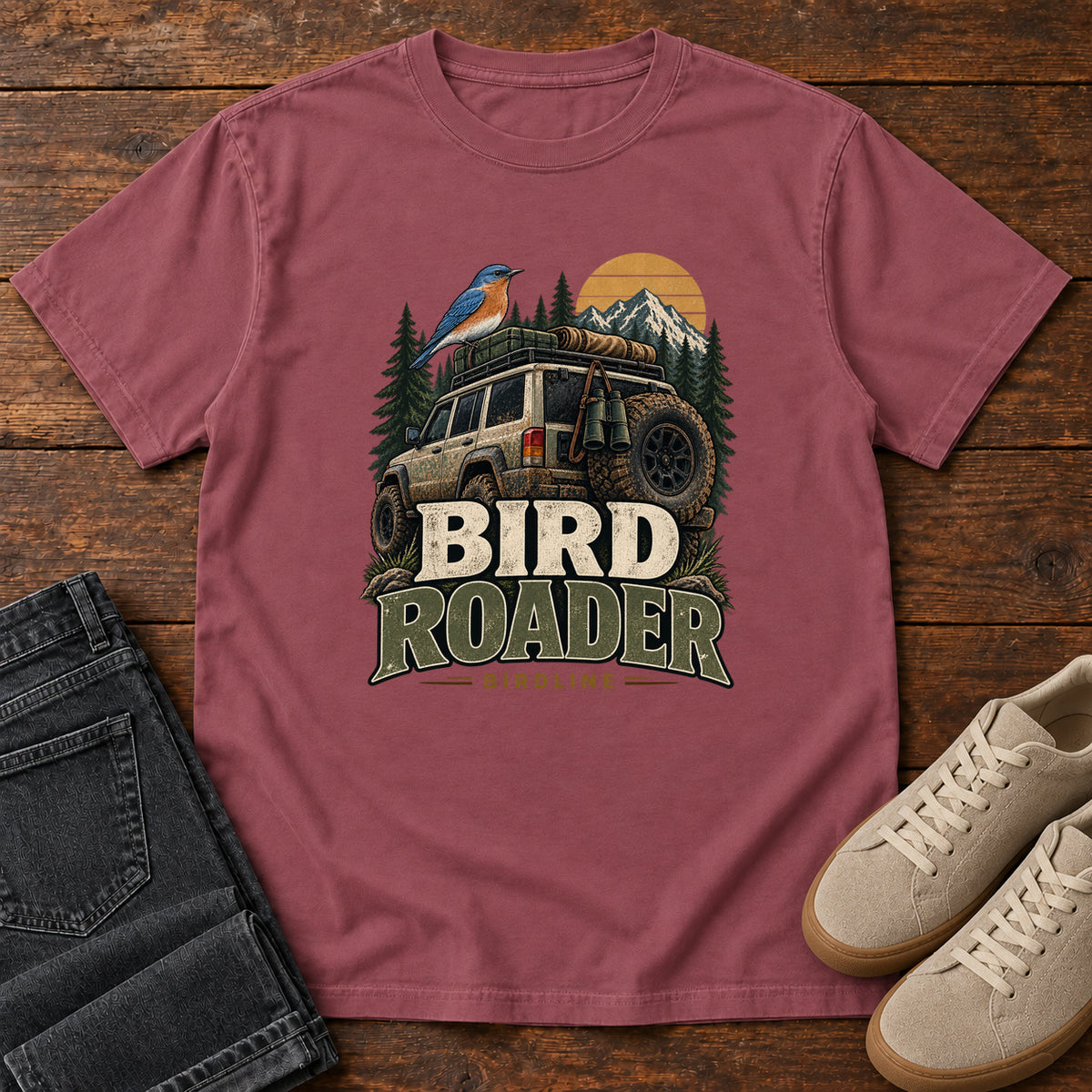 Brick Bird Roader Tailgate T-Shirt with a bird on an off-road vehicle beneath mountains and a golden sun; front flat lay on wood.