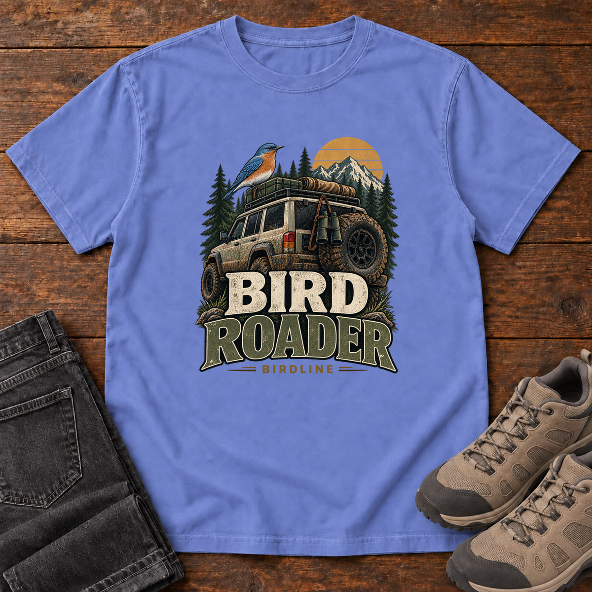 Flo Blue Bird Roader Tailgate T-Shirt with a bird on an off-road vehicle beneath mountains and a golden sun; front flat lay on wood.