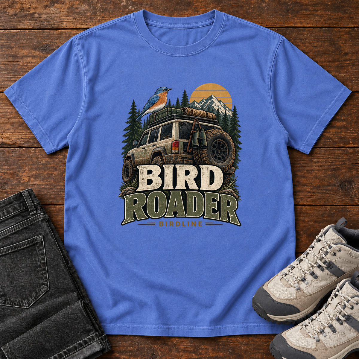 Mystic Blue Bird Roader Tailgate T-Shirt with a bird on an off-road vehicle beneath mountains and a golden sun; front flat lay on wood.