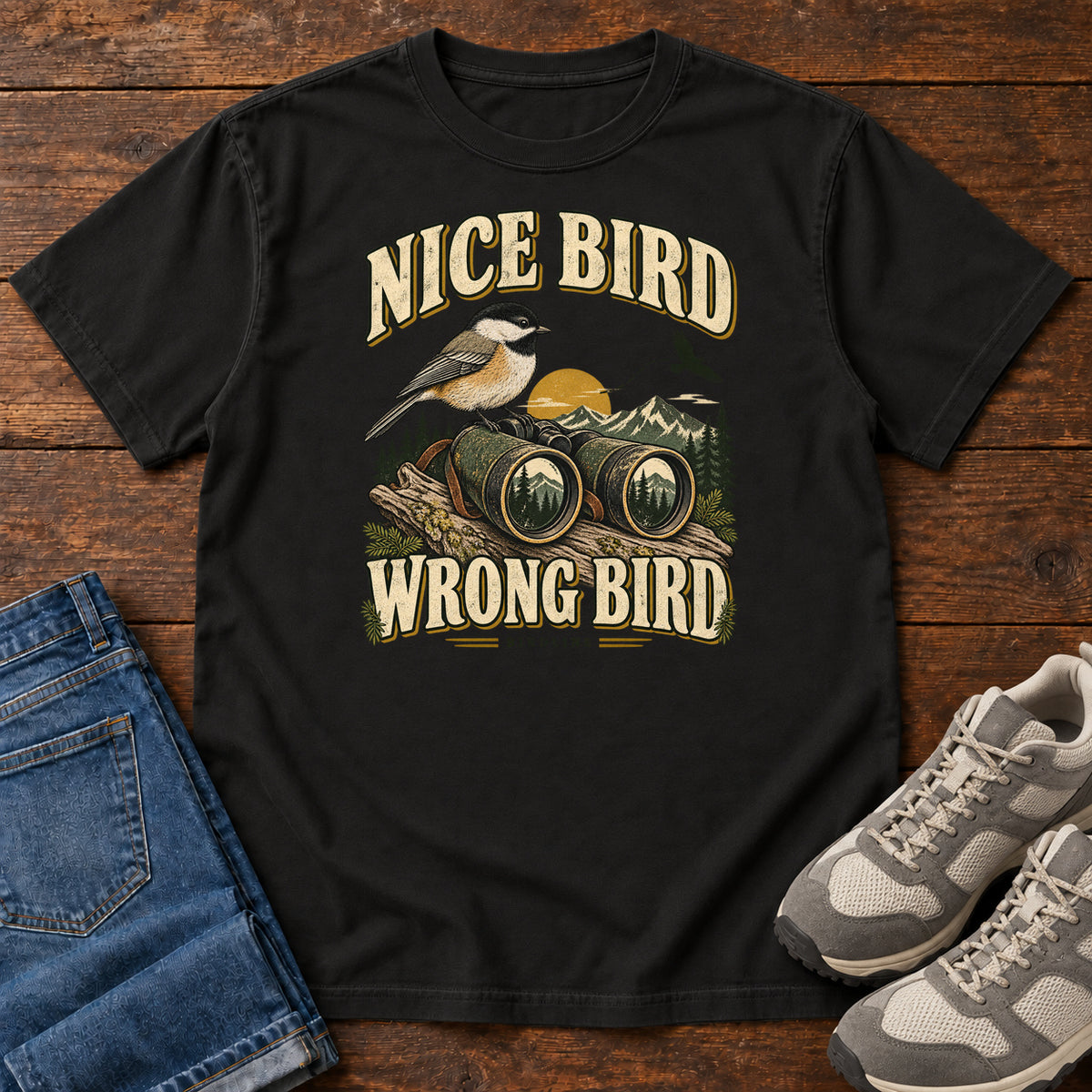 Black Nice Bird. Wrong Bird. T-Shirt with a small black-capped bird on binoculars against mountains and a golden sun; front flat lay on wood.