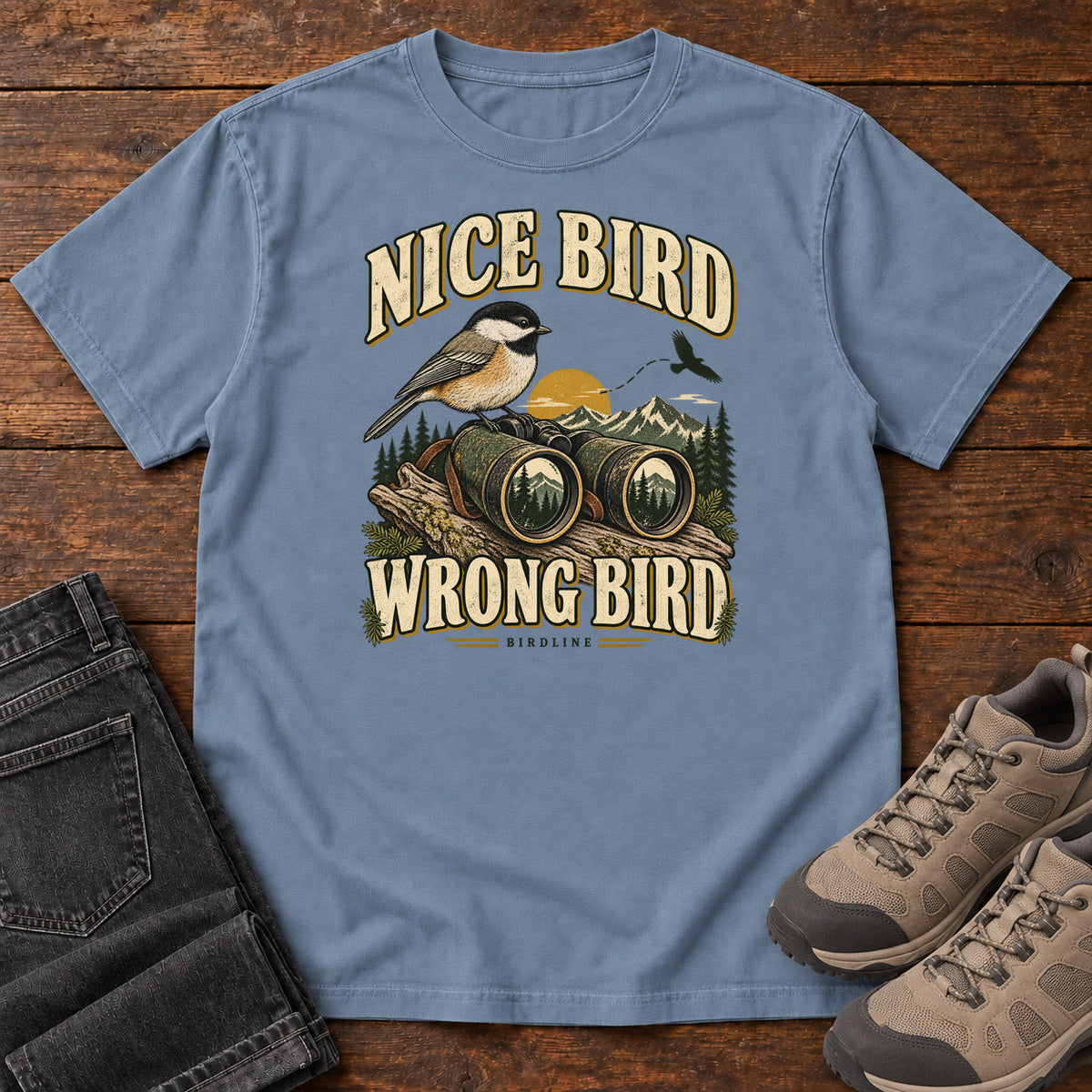 Blue Jean Nice Bird. Wrong Bird. T-Shirt with a small black-capped bird on binoculars against mountains and a golden sun; front flat lay on wood.
