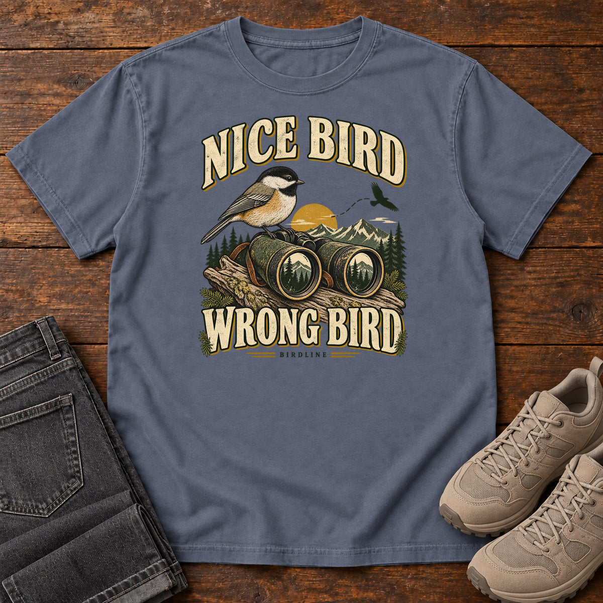 Denim Nice Bird. Wrong Bird. T-Shirt with a small black-capped bird on binoculars against mountains and a golden sun; front flat lay on wood.