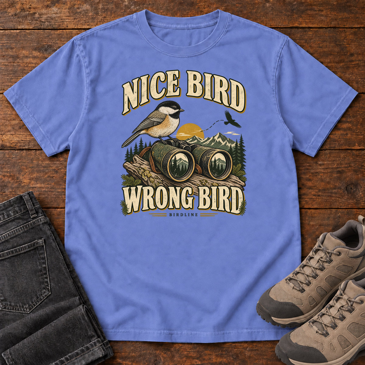 Flo Blue Nice Bird. Wrong Bird. T-Shirt with a small black-capped bird on binoculars against mountains and a golden sun; front flat lay on wood.