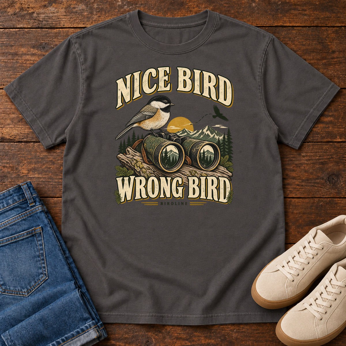 Graphite Nice Bird. Wrong Bird. T-Shirt with a small black-capped bird on binoculars against mountains and a golden sun; front flat lay on wood.