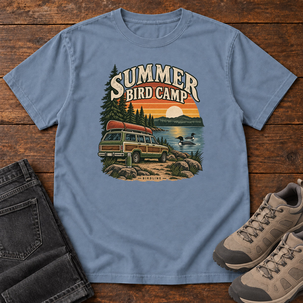 Summer Bird Camp T-Shirt in Blue Jean, front laid flat on wood, featuring a canoe-topped vehicle beside a lake with swimming birds, pine trees and a striped sunset.