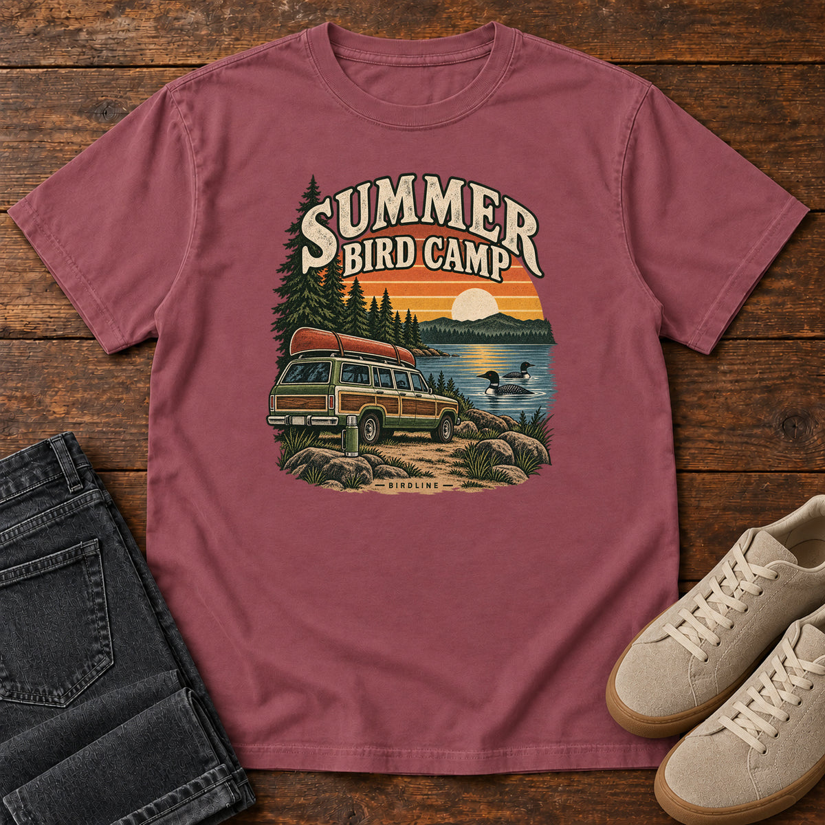 Summer Bird Camp T-Shirt in Brick, front laid flat on wood, featuring a canoe-topped vehicle beside a lake with swimming birds, pine trees and a striped sunset.