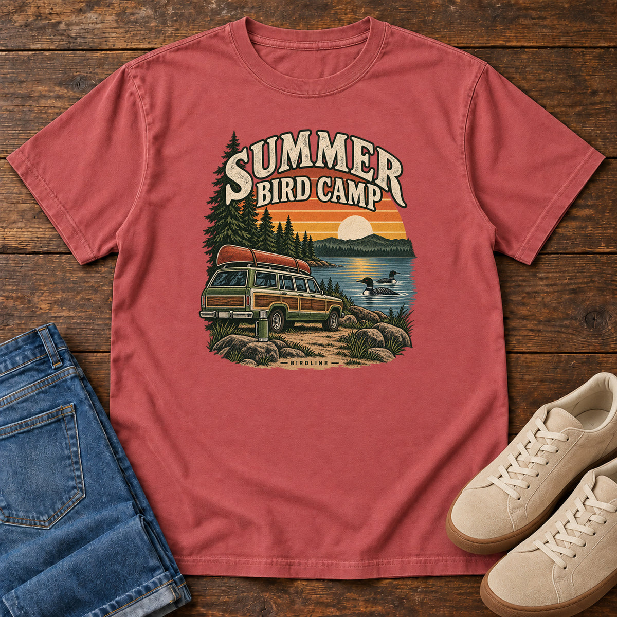 Summer Bird Camp T-Shirt in Crimson, front laid flat on wood, featuring a canoe-topped vehicle beside a lake with swimming birds, pine trees and a striped sunset.