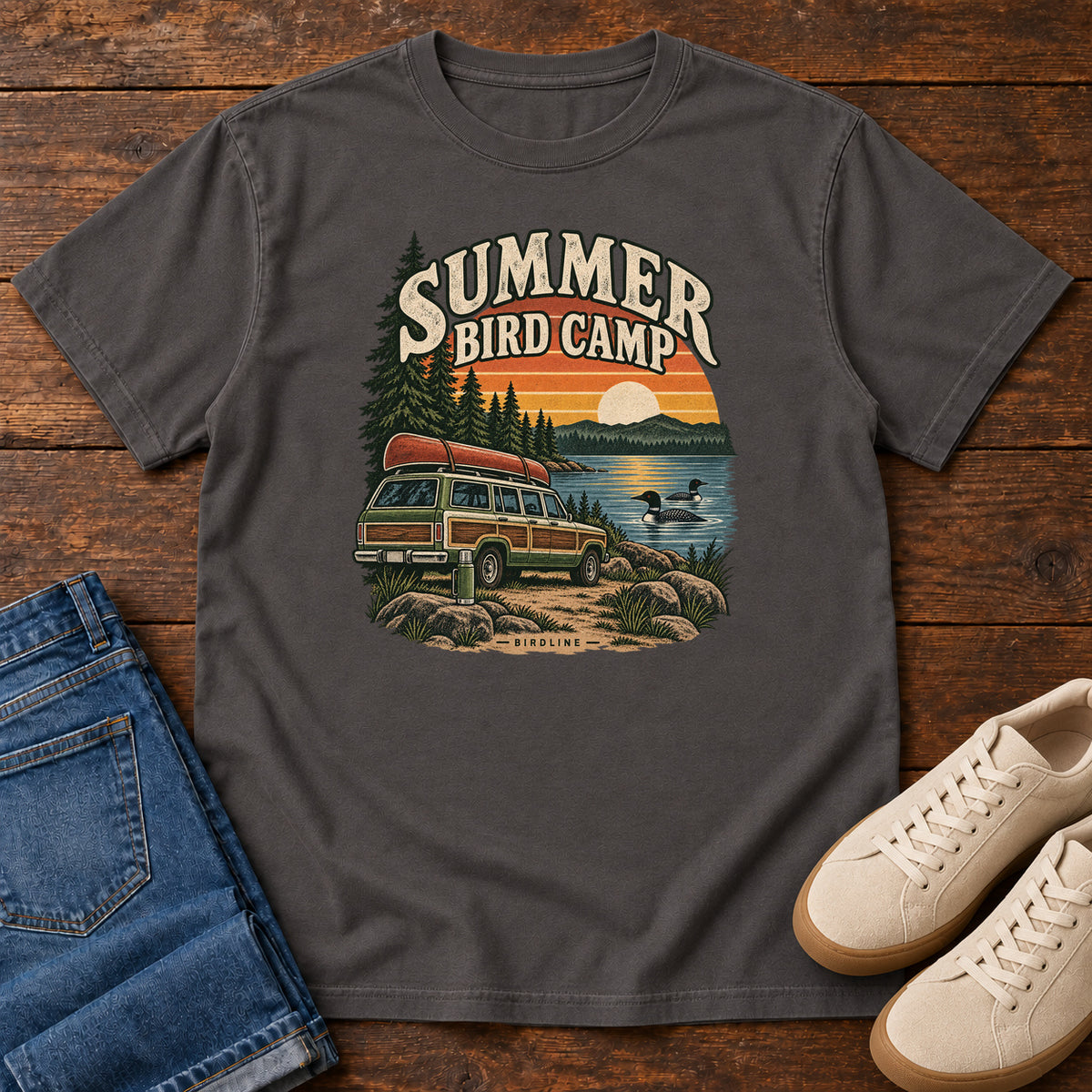 Summer Bird Camp T-Shirt in Graphite, front laid flat on wood, featuring a canoe-topped vehicle beside a lake with swimming birds, pine trees and a striped sunset.