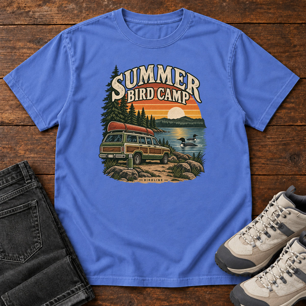 Summer Bird Camp T-Shirt in Mystic Blue, front laid flat on wood, featuring a canoe-topped vehicle beside a lake with swimming birds, pine trees and a striped sunset.
