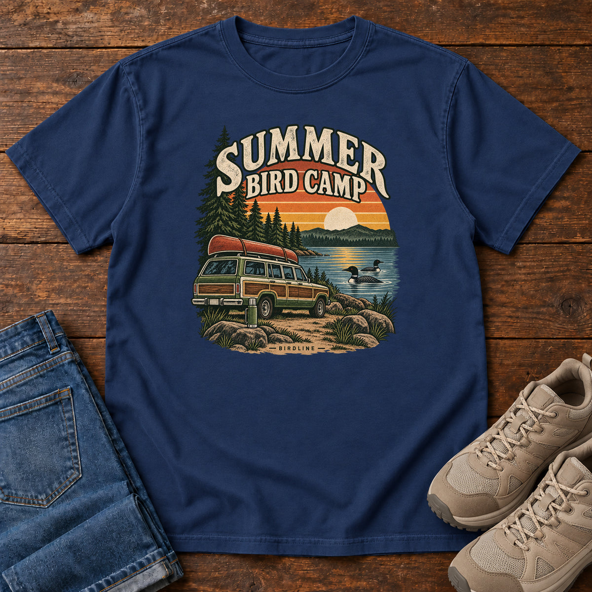 Summer Bird Camp T-Shirt in True Navy, front laid flat on wood, featuring a canoe-topped vehicle beside a lake with swimming birds, pine trees and a striped sunset.