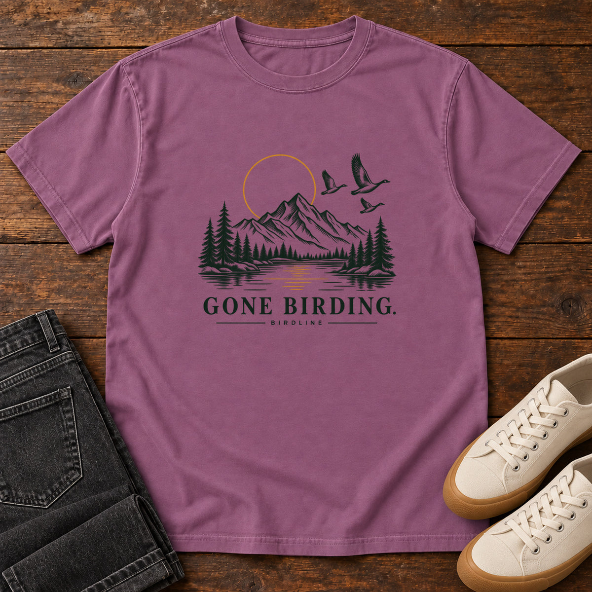 Berry Gone Birding T-Shirt with mountains, evergreen trees, flying birds and an outlined golden sun, front flat lay on wood.