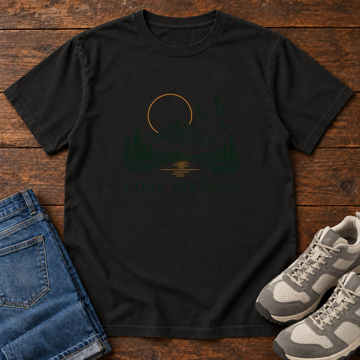 Black Gone Birding T-Shirt with mountains, evergreen trees, flying birds and an outlined golden sun, front flat lay on wood.