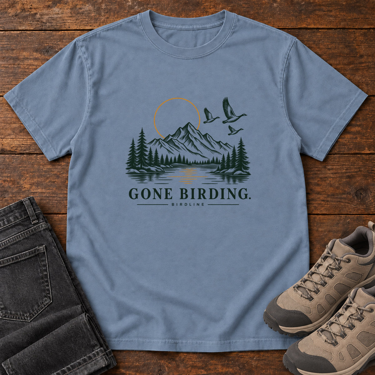 Blue Jean Gone Birding T-Shirt with mountains, evergreen trees, flying birds and an outlined golden sun, front flat lay on wood.