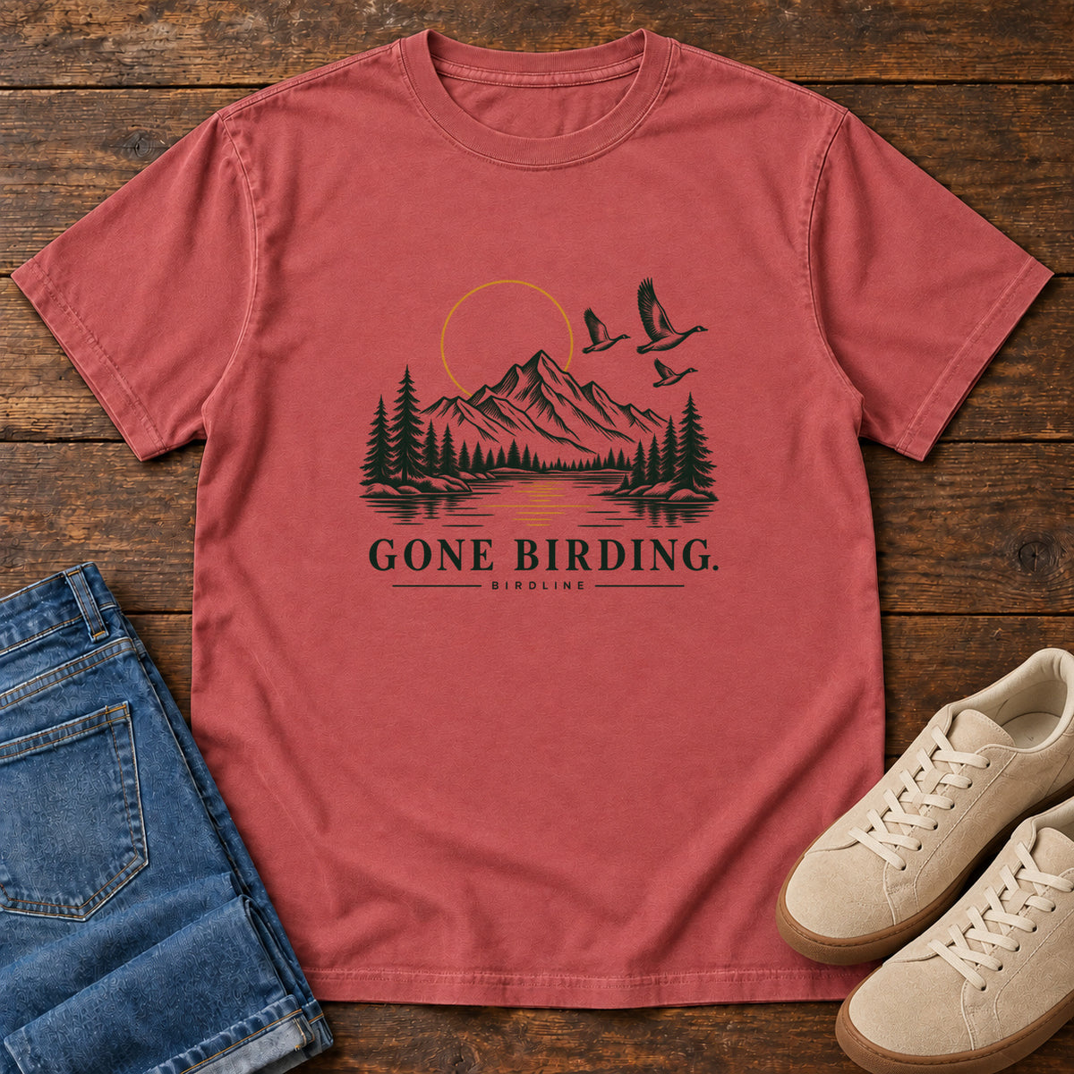 Crimson Gone Birding T-Shirt with mountains, evergreen trees, flying birds and an outlined golden sun, front flat lay on wood.
