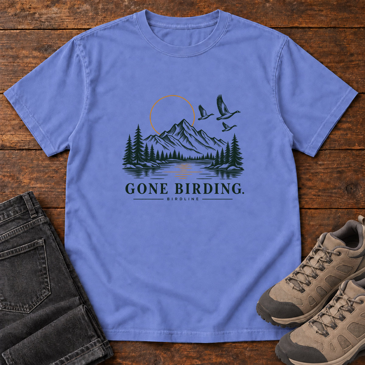 Flo Blue Gone Birding T-Shirt with mountains, evergreen trees, flying birds and an outlined golden sun, front flat lay on wood.