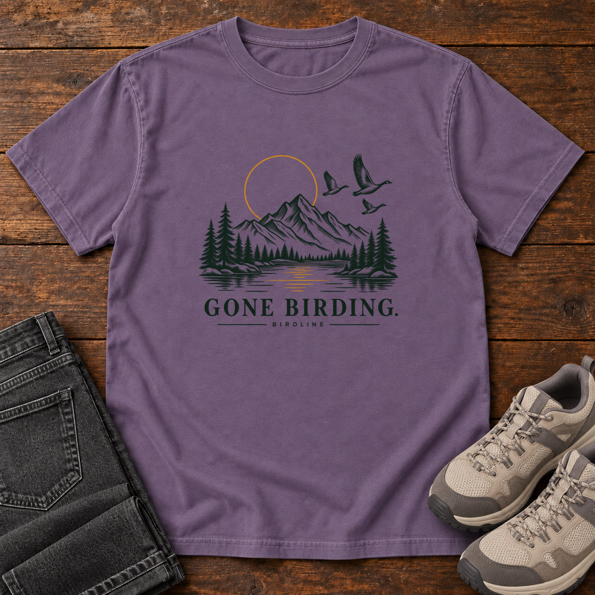 Grape Gone Birding T-Shirt with mountains, evergreen trees, flying birds and an outlined golden sun, front flat lay on wood.