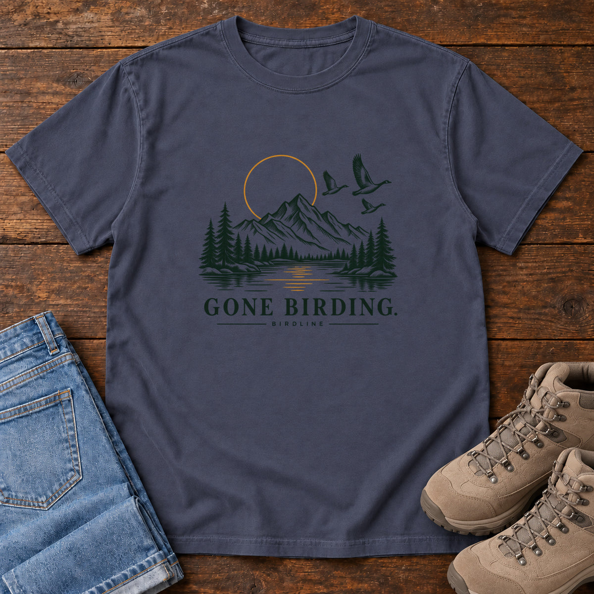 Navy Gone Birding T-Shirt with mountains, evergreen trees, flying birds and an outlined golden sun, front flat lay on wood.
