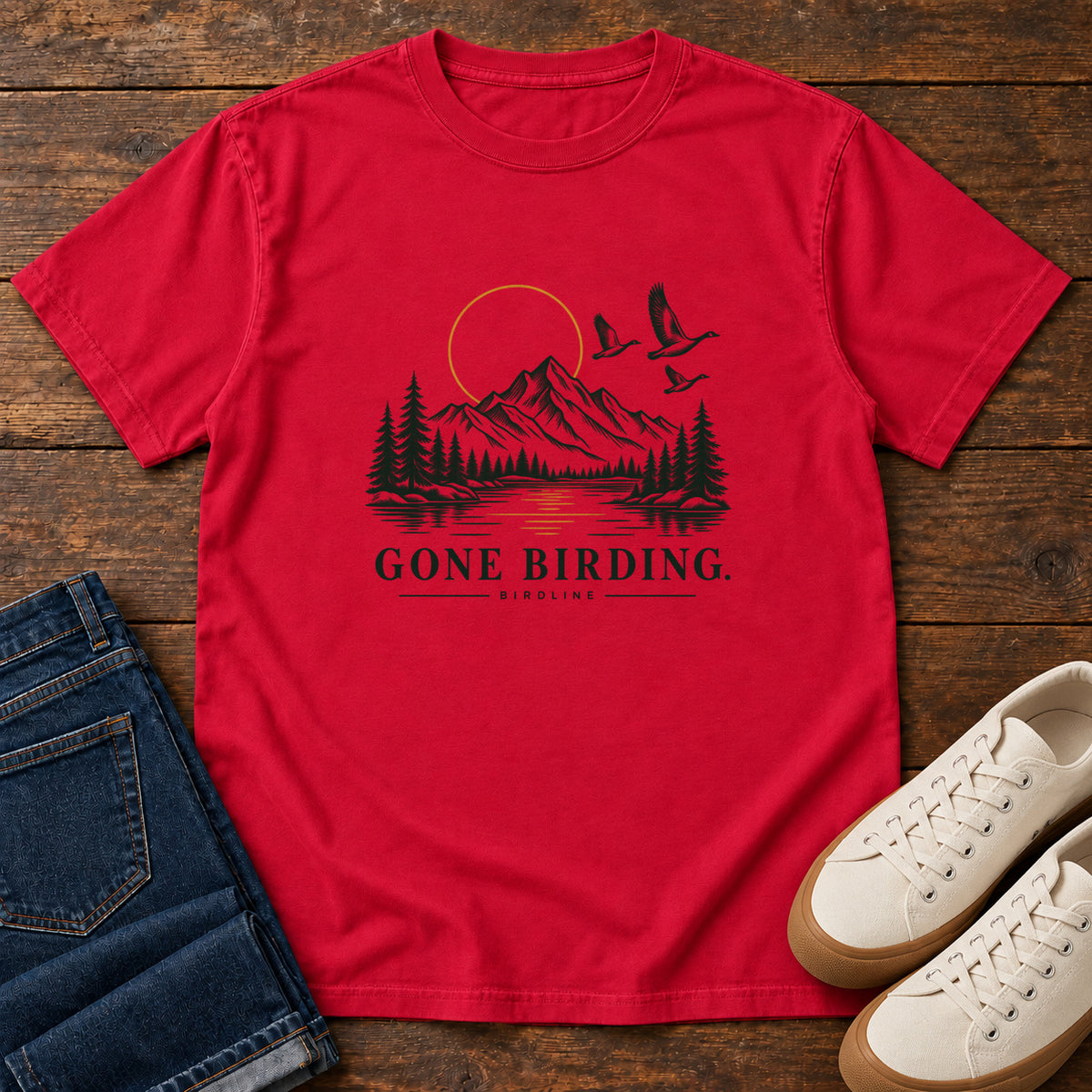 Red Gone Birding T-Shirt with mountains, evergreen trees, flying birds and an outlined golden sun, front flat lay on wood.