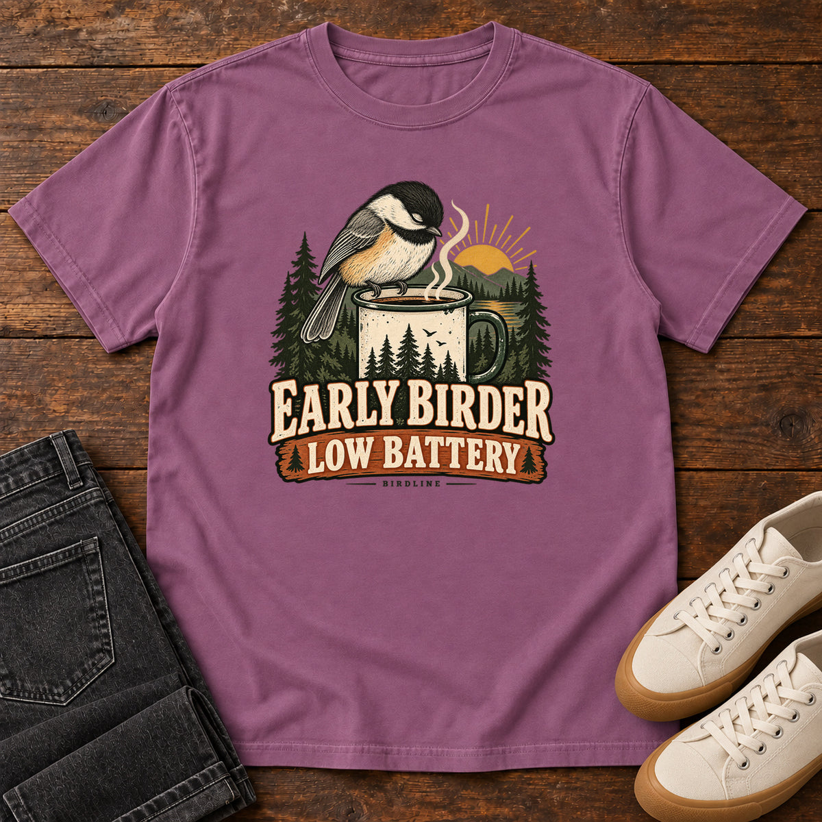 Berry Early Birder. Low Battery. T-Shirt with a bird perched on a steaming mug, evergreens and a rising sun; front flat lay on wood.