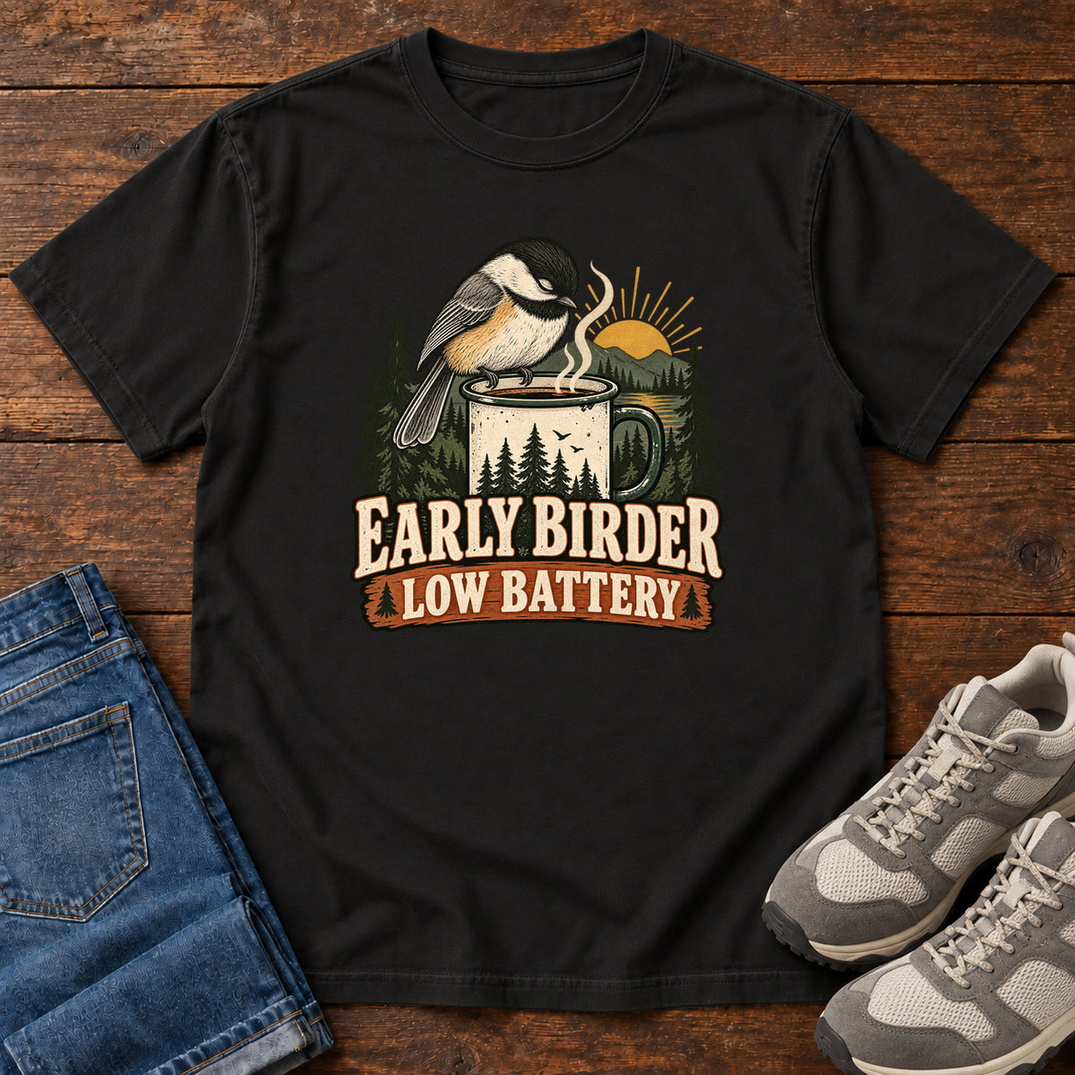 Black Early Birder. Low Battery. T-Shirt with a bird perched on a steaming mug, evergreens and a rising sun; front flat lay on wood.