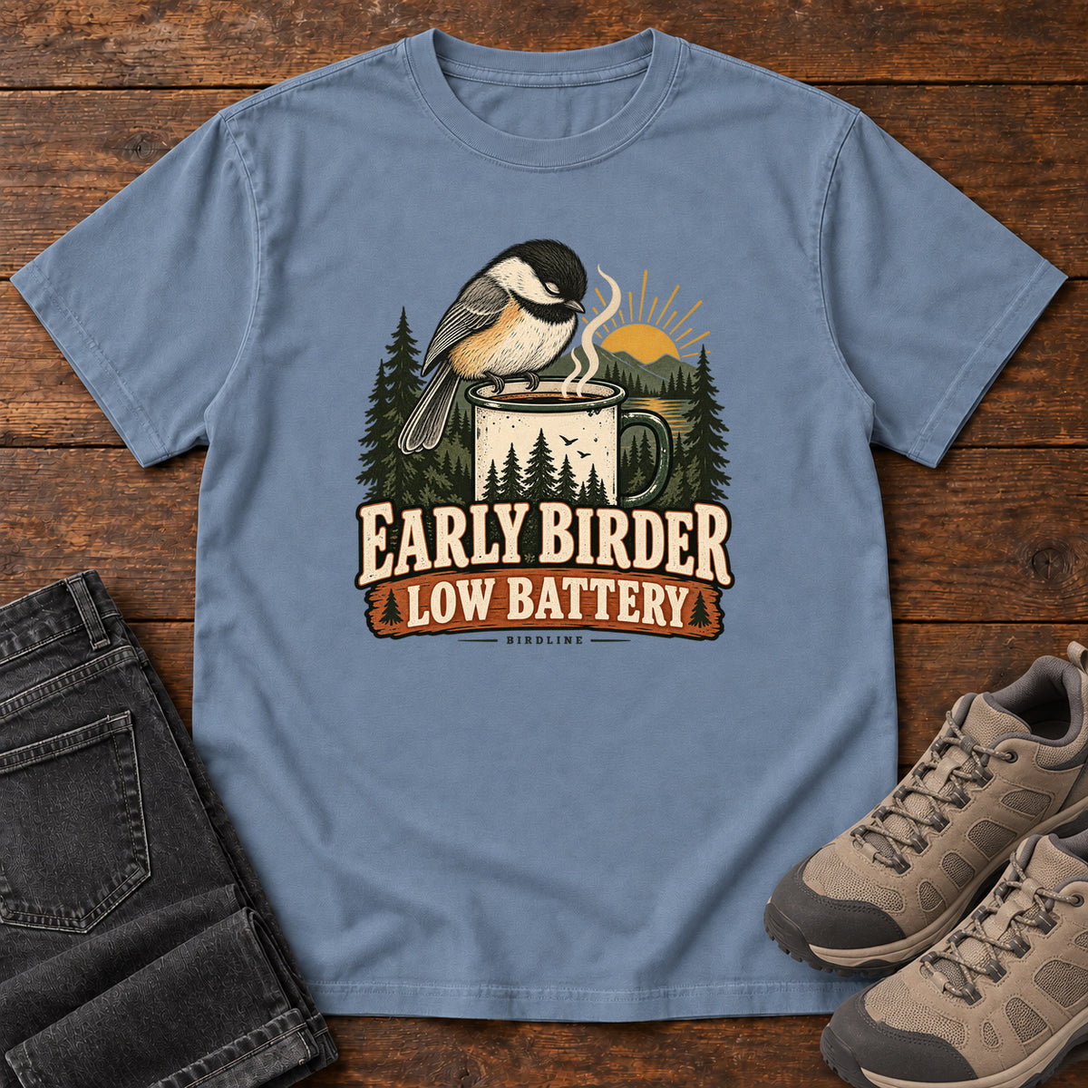 Blue Jean Early Birder. Low Battery. T-Shirt with a bird perched on a steaming mug, evergreens and a rising sun; front flat lay on wood.
