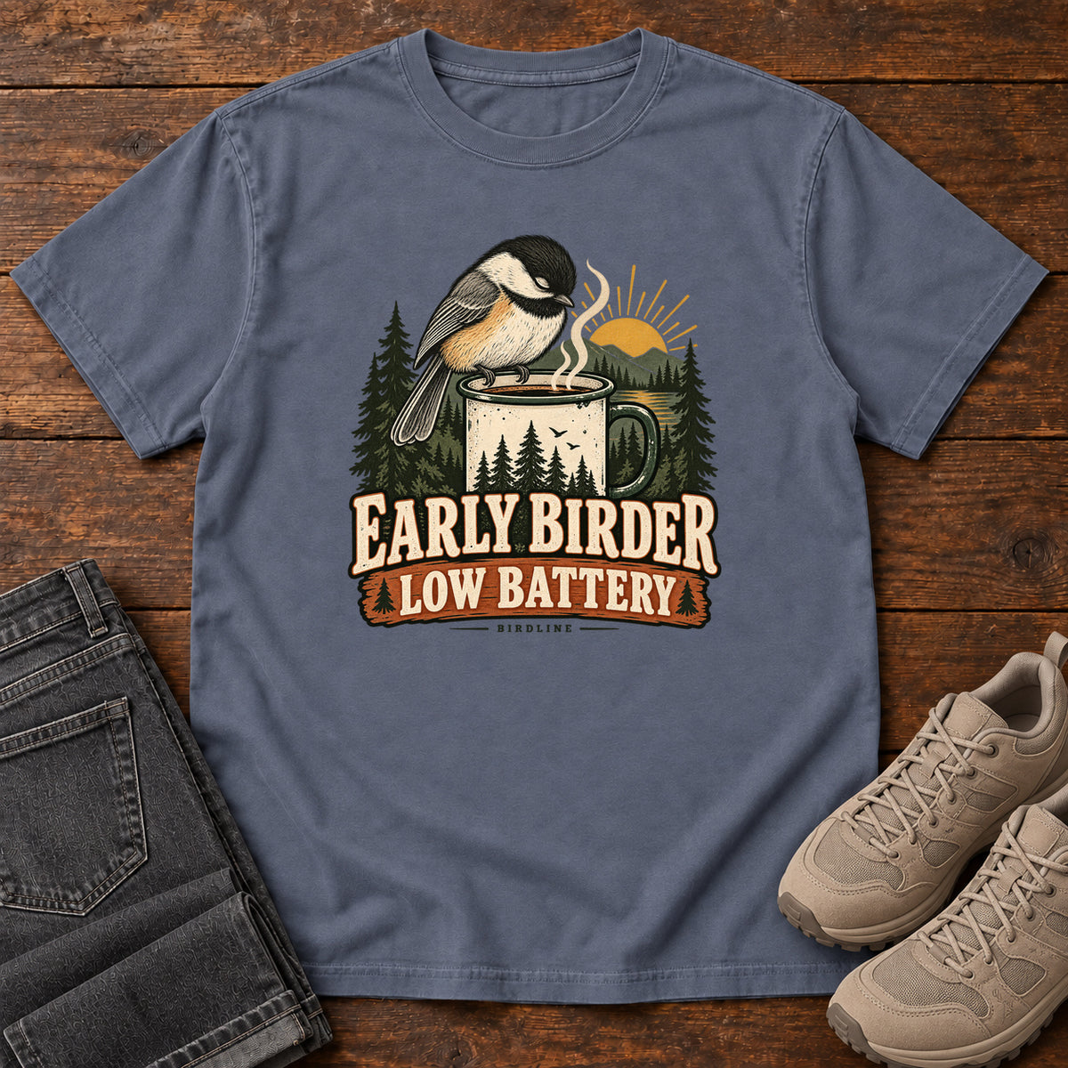 Denim Early Birder. Low Battery. T-Shirt with a bird perched on a steaming mug, evergreens and a rising sun; front flat lay on wood.