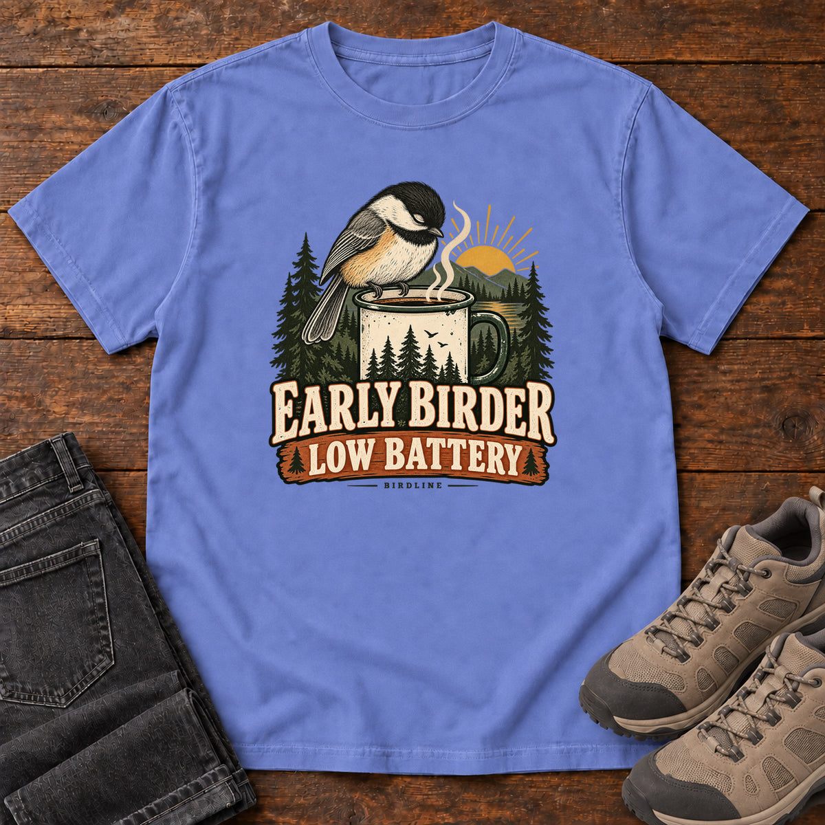 Flo Blue Early Birder. Low Battery. T-Shirt with a bird perched on a steaming mug, evergreens and a rising sun; front flat lay on wood.