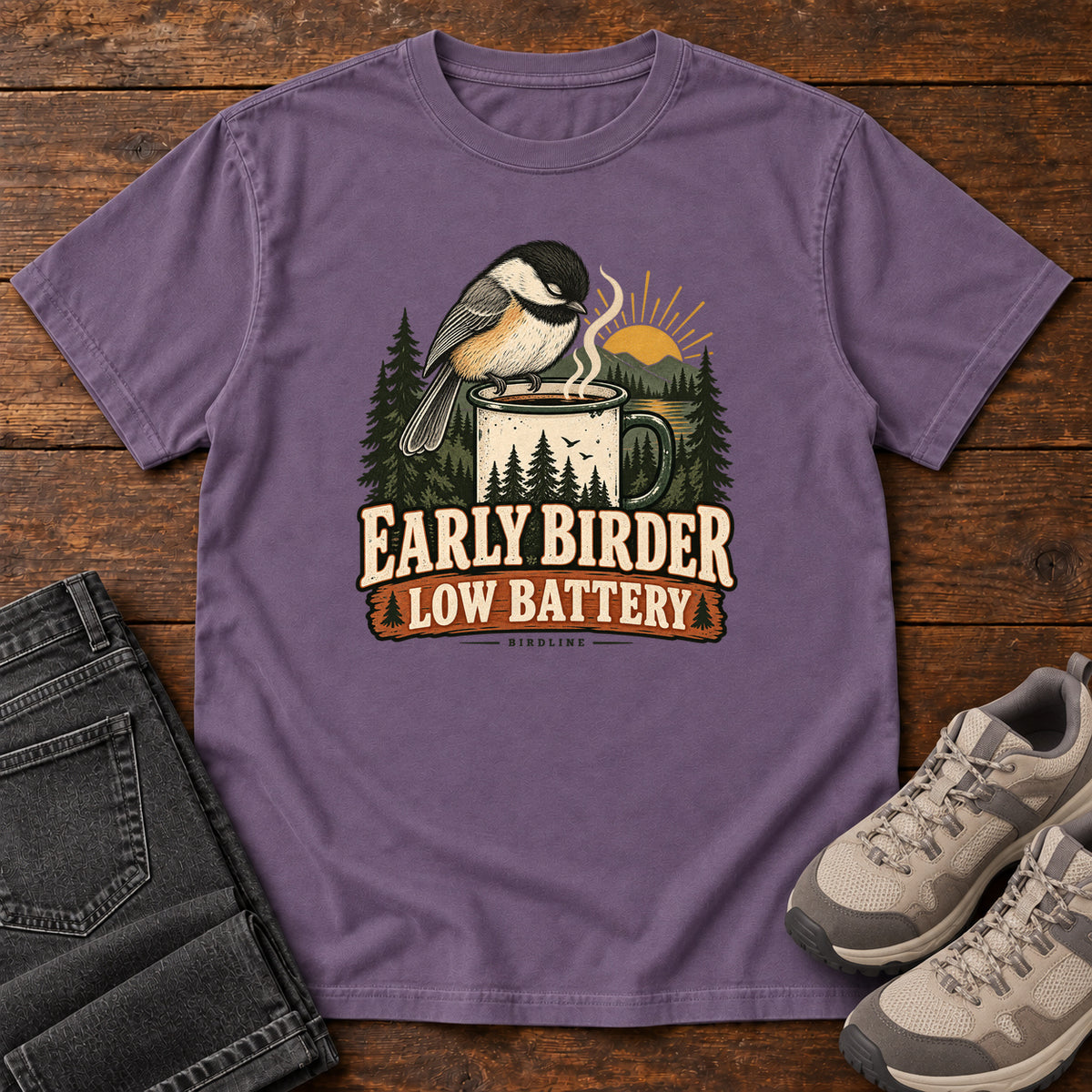 Grape Early Birder. Low Battery. T-Shirt with a bird perched on a steaming mug, evergreens and a rising sun; front flat lay on wood.
