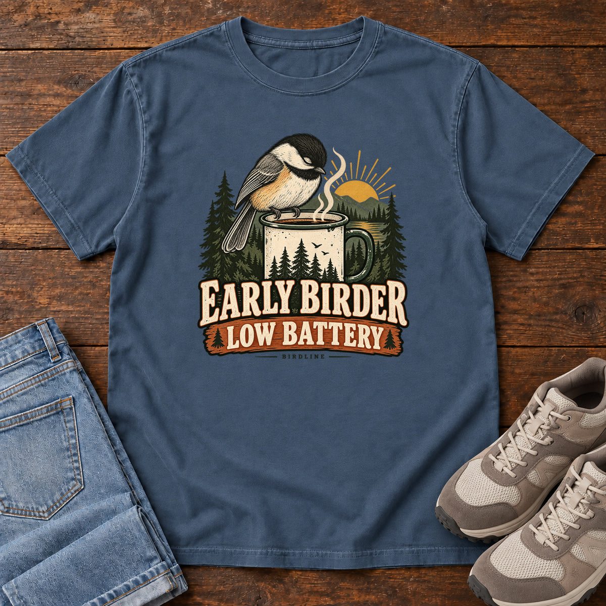 Midnight Early Birder. Low Battery. T-Shirt with a bird perched on a steaming mug, evergreens and a rising sun; front flat lay on wood.