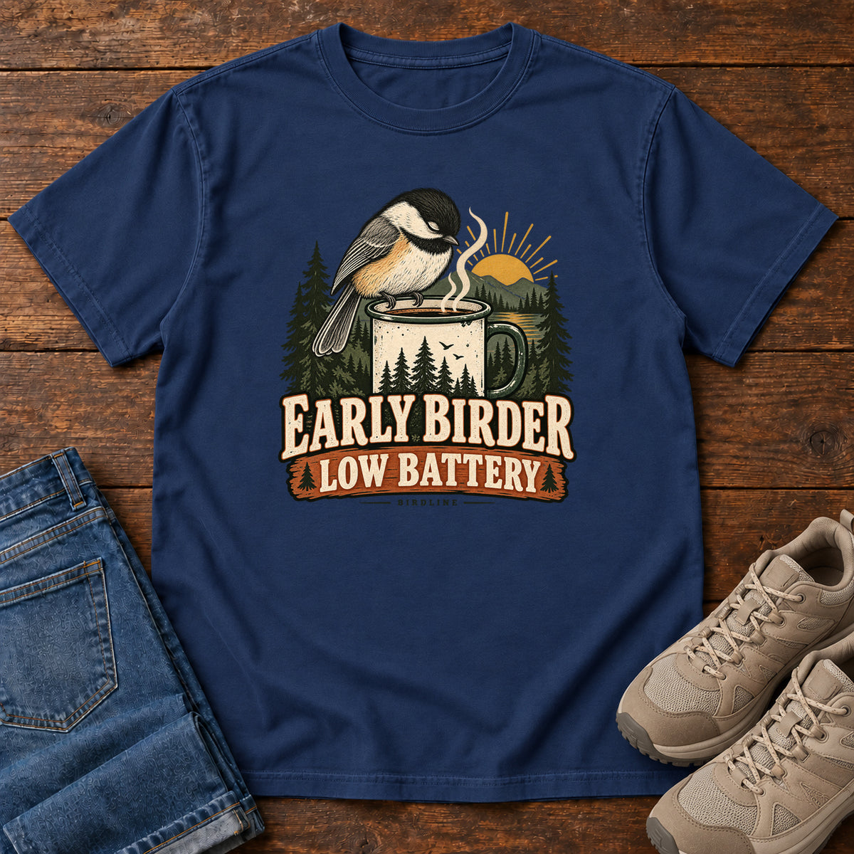 True Navy Early Birder. Low Battery. T-Shirt with a bird perched on a steaming mug, evergreens and a rising sun; front flat lay on wood.