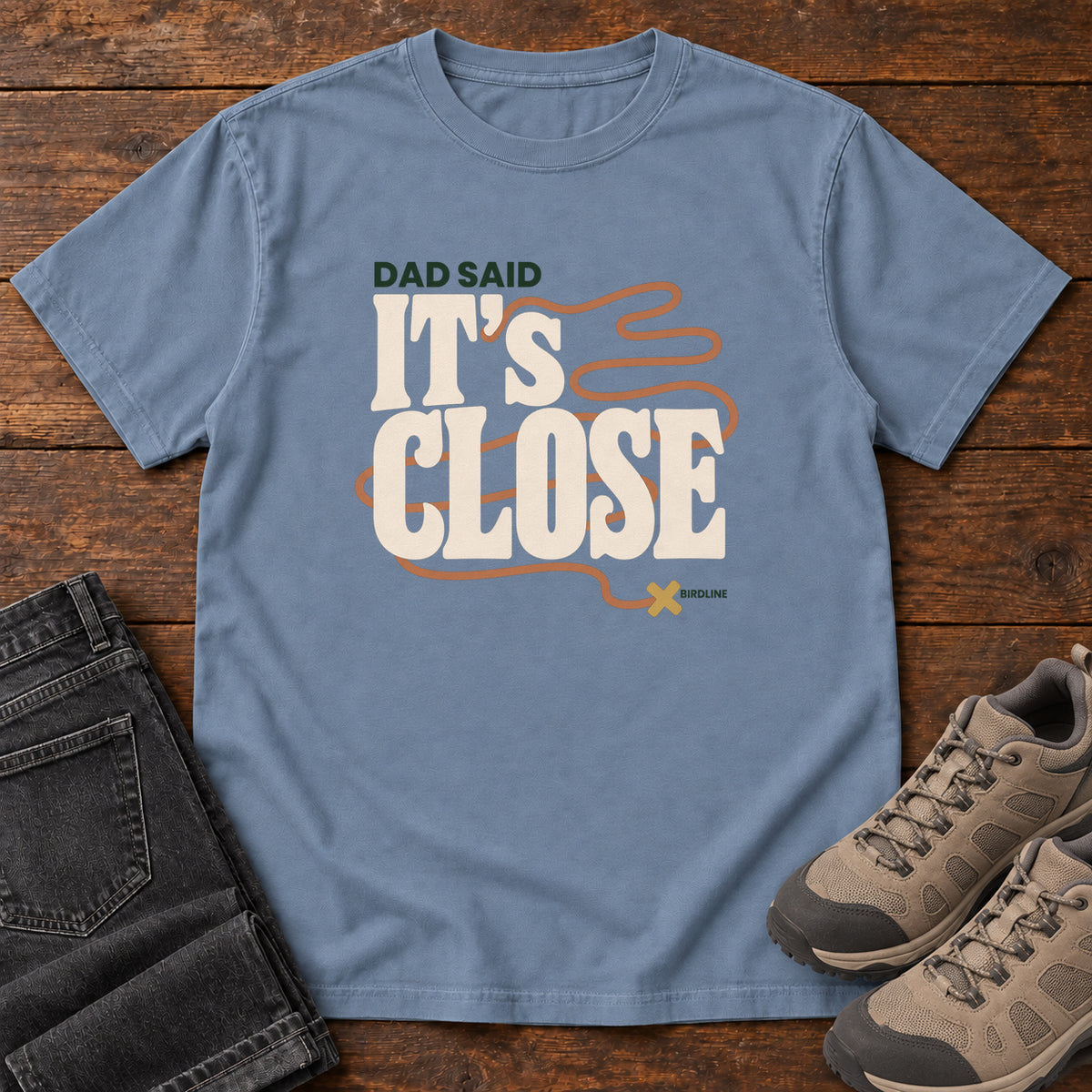 Blue Jean Dad Said It's Close T-Shirt with bold cream lettering wrapped by a winding orange route ending at an X; front flat lay on wood.