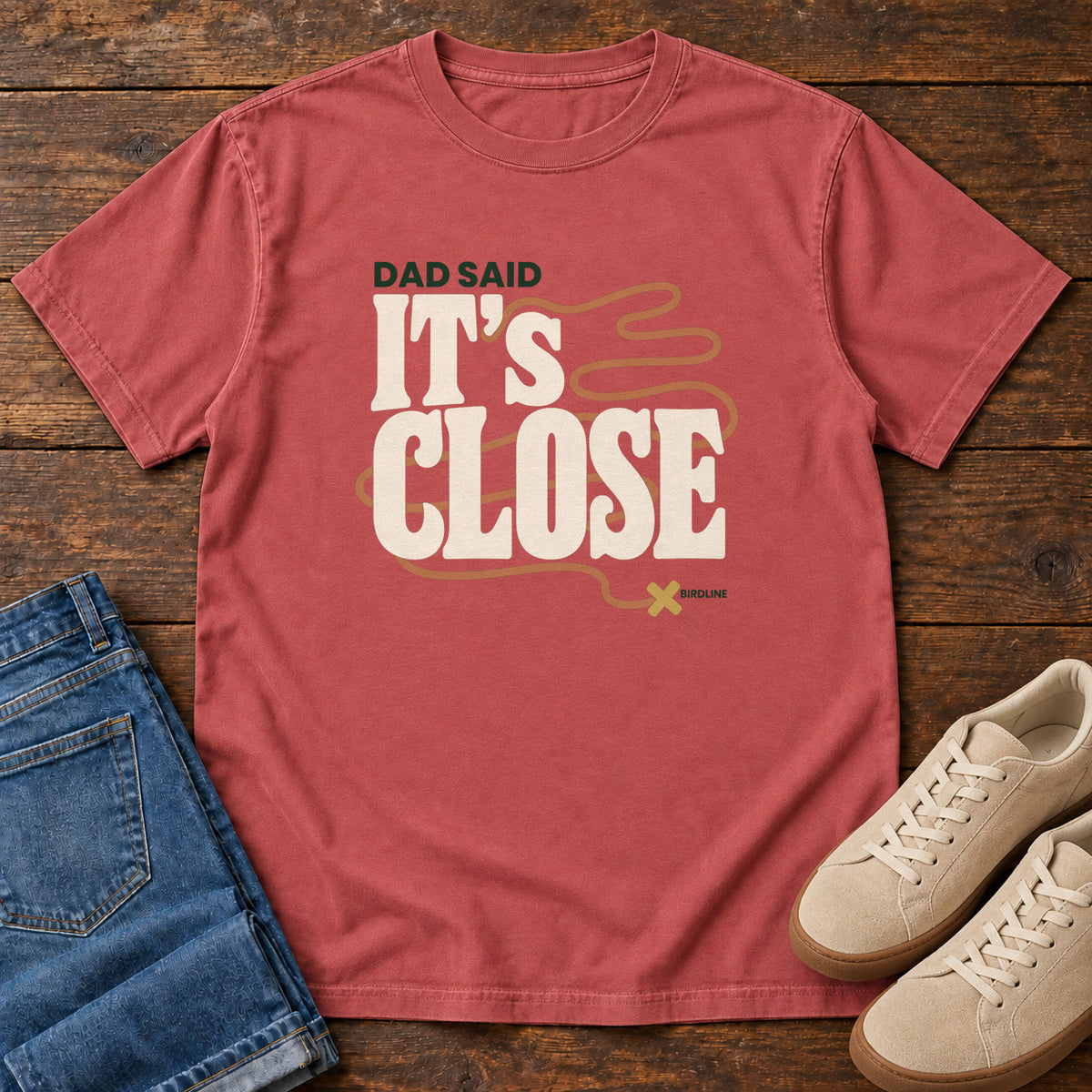 Crimson Dad Said It's Close T-Shirt with bold cream lettering wrapped by a winding orange route ending at an X; front flat lay on wood.
