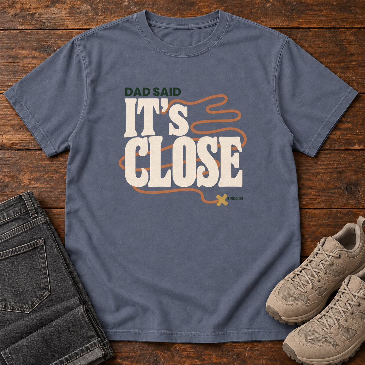 Denim Dad Said It's Close T-Shirt with bold cream lettering wrapped by a winding orange route ending at an X; front flat lay on wood.