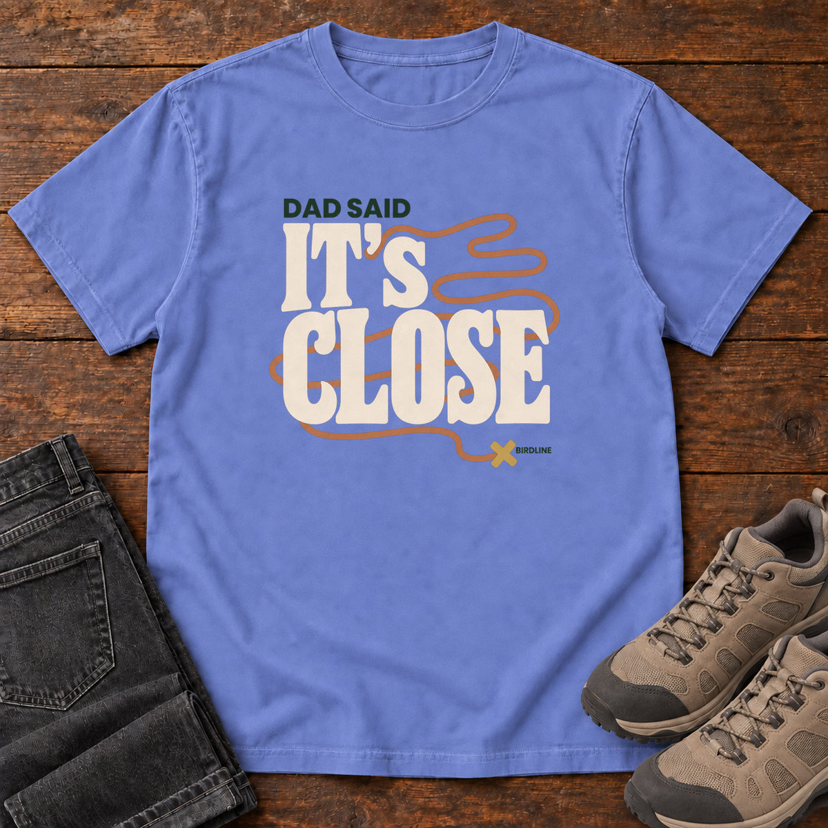 Flo Blue Dad Said It's Close T-Shirt with bold cream lettering wrapped by a winding orange route ending at an X; front flat lay on wood.