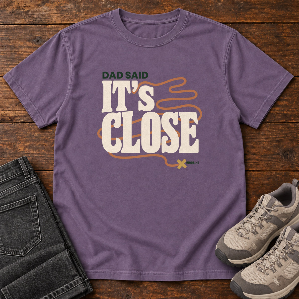 Grape Dad Said It's Close T-Shirt with bold cream lettering wrapped by a winding orange route ending at an X; front flat lay on wood.