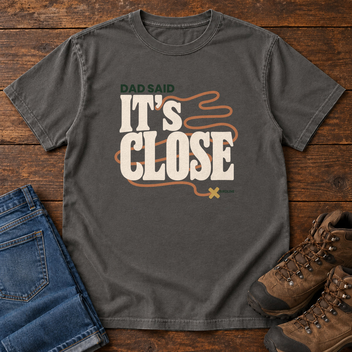 Pepper Dad Said It's Close T-Shirt with bold cream lettering wrapped by a winding orange route ending at an X; front flat lay on wood.